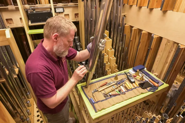 An organ builder voicing a metal pipe at the workbench, surrounded by racked pipework and hand tools
